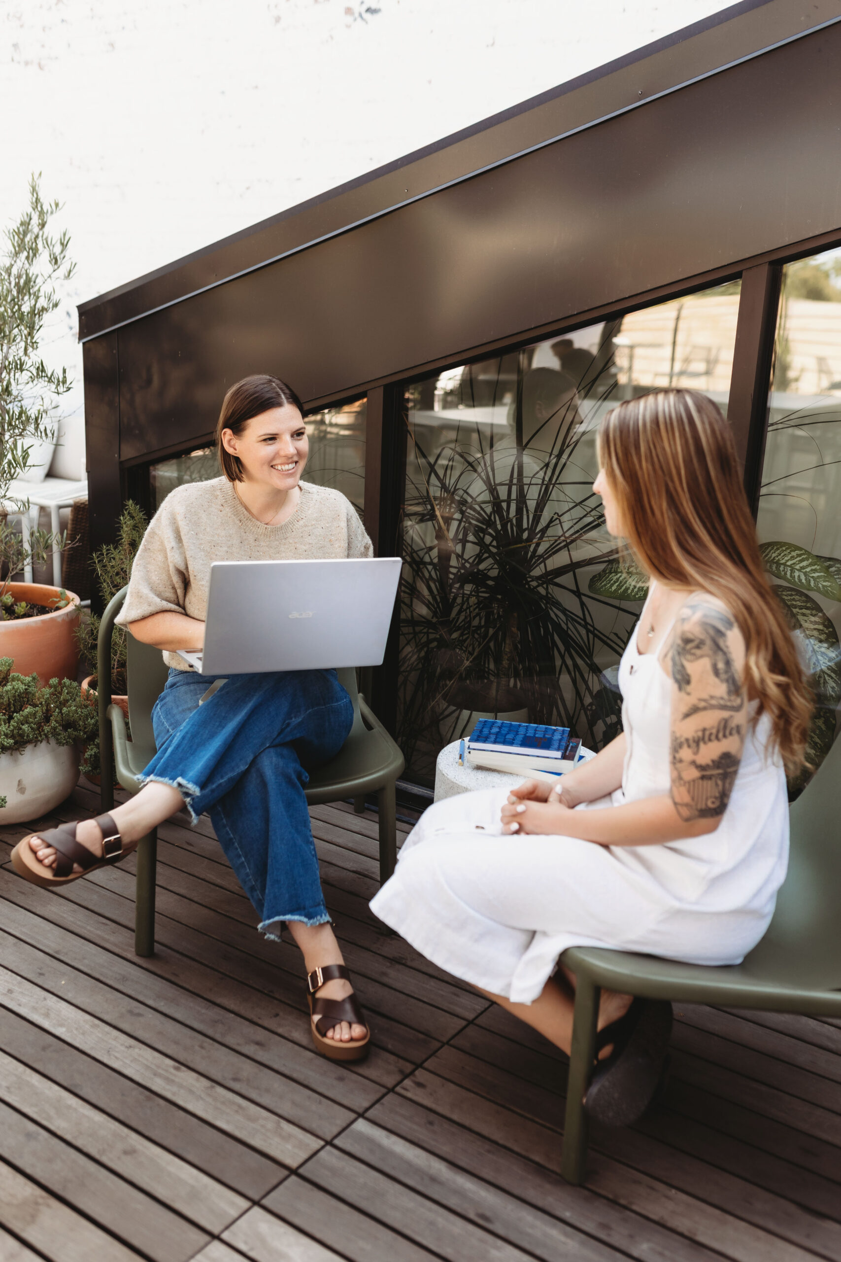 Two women sit on a wooden patio, engaged in conversation. One is holding a laptop and wearing a casual outfit with jeans, while the other, dressed in a white dress, has a visible arm tattoo. The setting is relaxed and sunlit, suggesting an informal outdoor meeting or friendly catch-up.