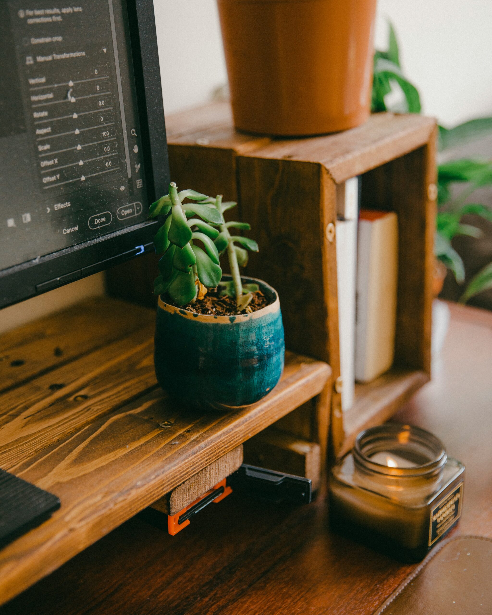 A close-up of a wooden desk setup with a computer monitor displaying editing software, a small potted succulent in a teal pot, stacked books, a candle, and a larger plant in a terracotta pot. The scene has a warm, earthy aesthetic and cozy, organized vibe.