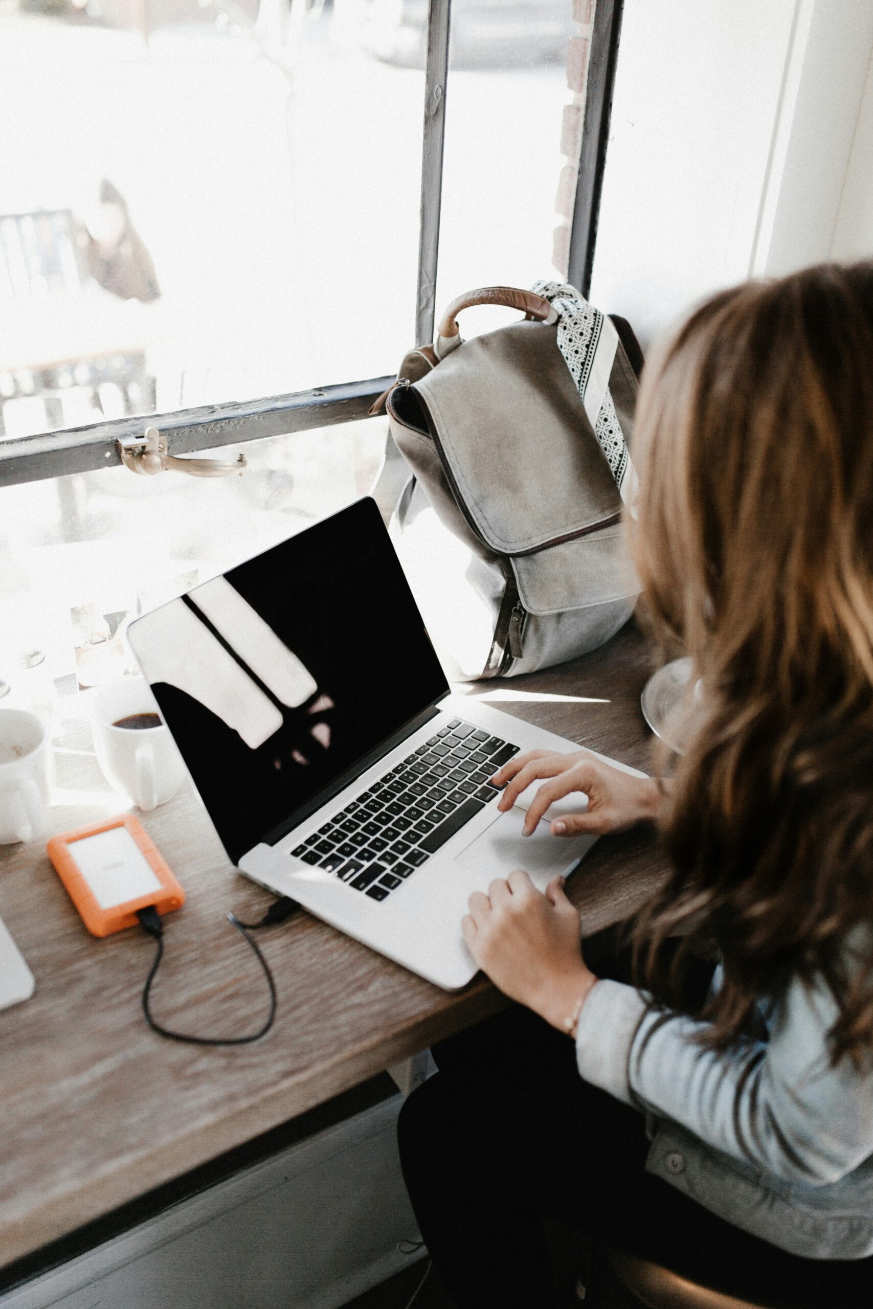 A woman with long hair is working on a laptop at a wooden table near a window, with a grey backpack, external hard drive, and coffee cup nearby. Natural light fills the space, suggesting a casual work environment, possibly a café or co-working spot.