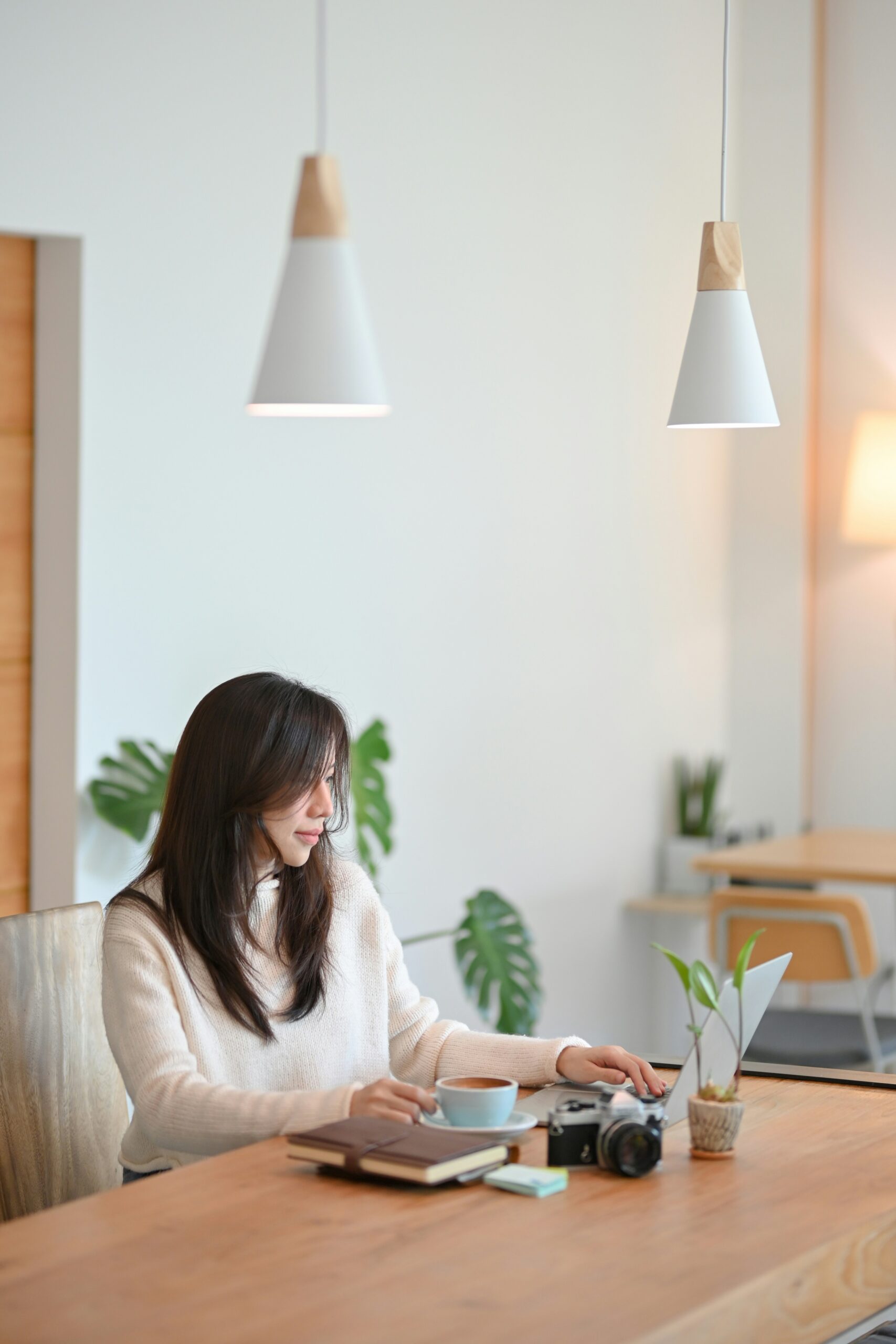A woman with long dark hair is working at a desk on a laptop in a bright, minimalist room with potted plants and soft lighting. She is wearing a cozy cream-colored sweater, and the setting appears calm and focused.