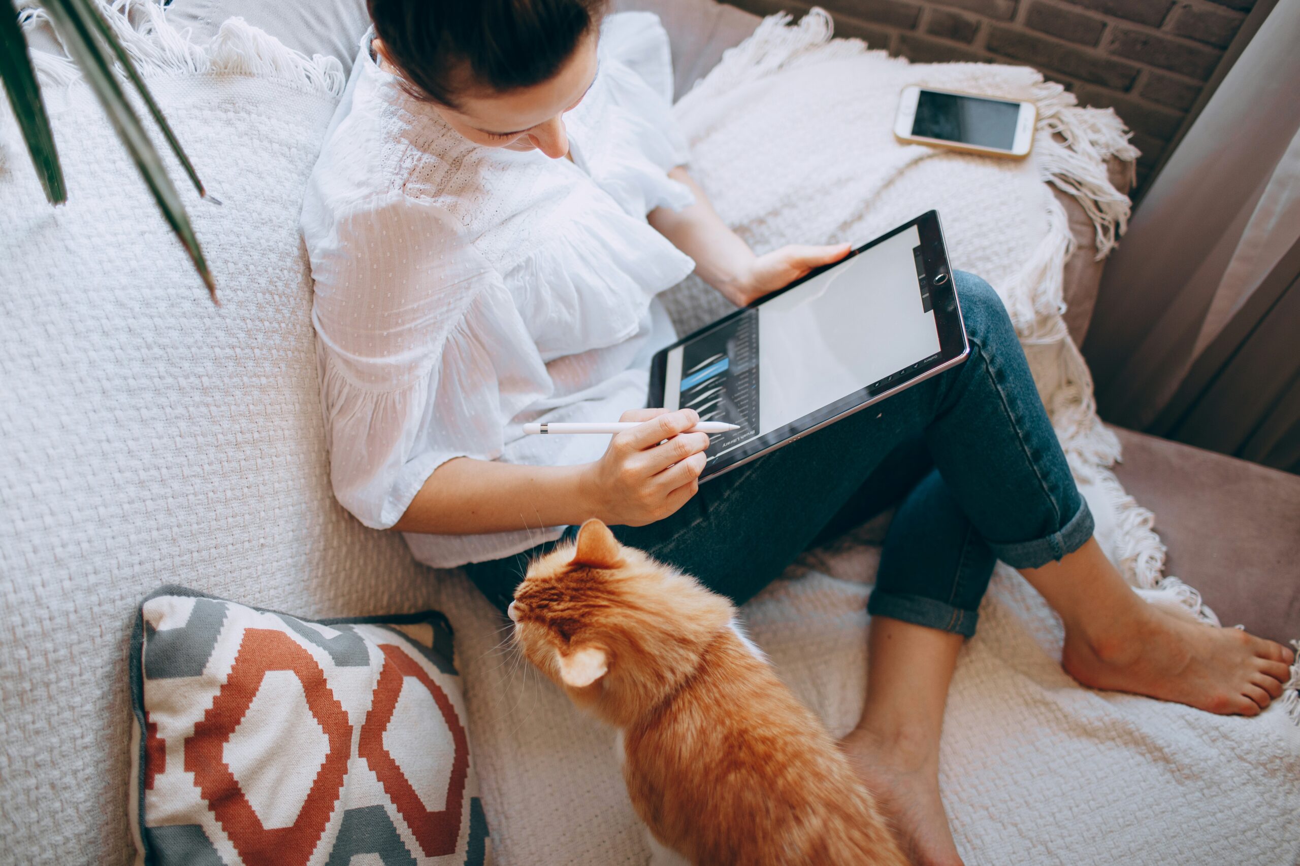 A woman sits cross-legged on a cozy couch using a tablet with a stylus while a ginger cat looks up at her. The setting feels warm and relaxed, blending productivity with comfort and a touch of companionship.