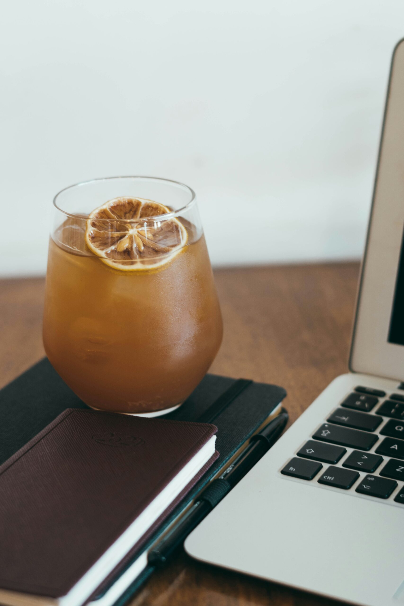 A glass of iced coffee or cold brew topped with a dried citrus slice sits on top of a closed notebook beside a laptop on a wooden desk. The scene suggests a relaxed work or study session with a refreshing drink.