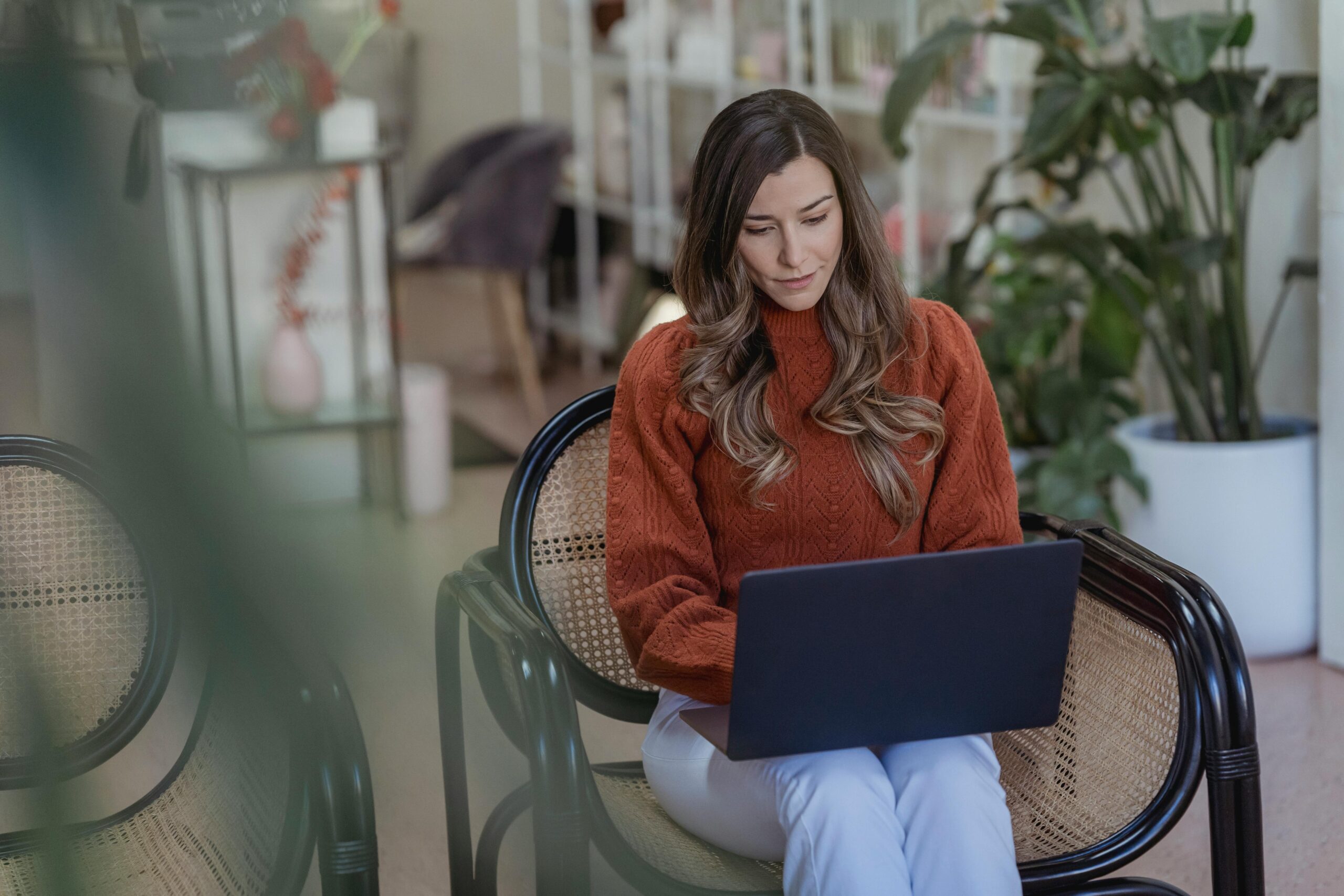 A woman in a rust-colored sweater sits on a woven-back chair, focused on her laptop in a cozy, plant-filled indoor setting. The background hints at a relaxed workspace or café environment with soft lighting and homey decor.