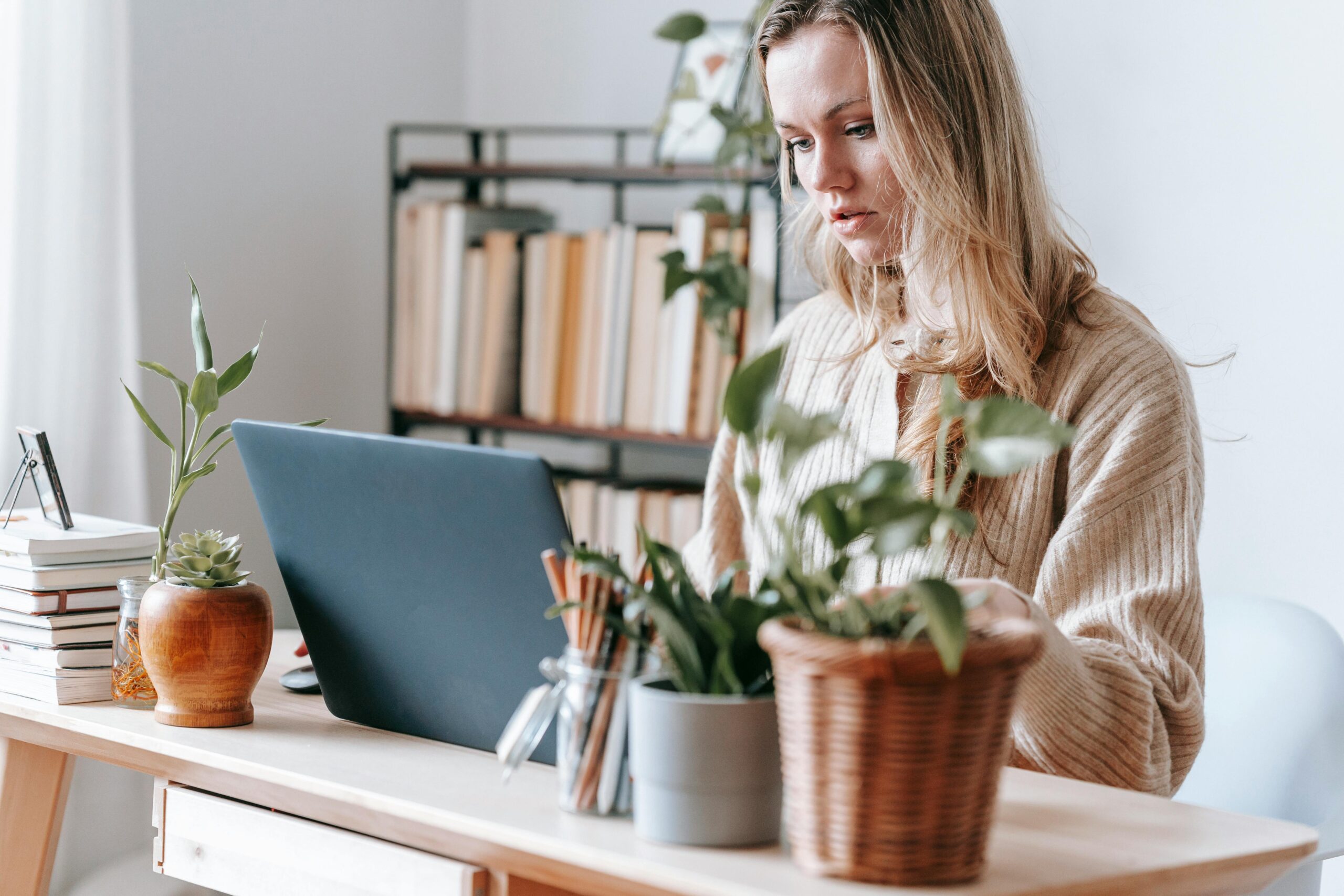 A blonde woman works at a desk with a laptop, surrounded by potted plants and stationery, with a bookshelf full of books in the background. The setting feels calm and homey, blending productivity with a touch of greenery.