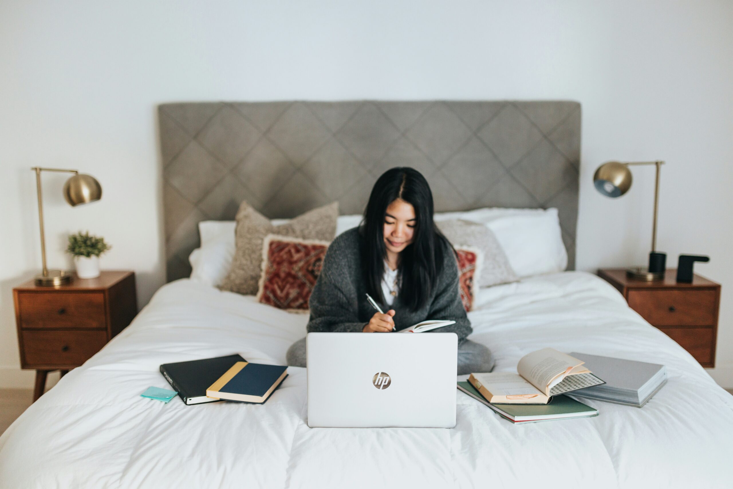 A woman sits on a neatly made bed with a laptop, notebook, and open books spread around her, appearing focused as she writes. The room features a tufted headboard, warm lighting, and a cozy, organized vibe—ideal for studying or remote work.